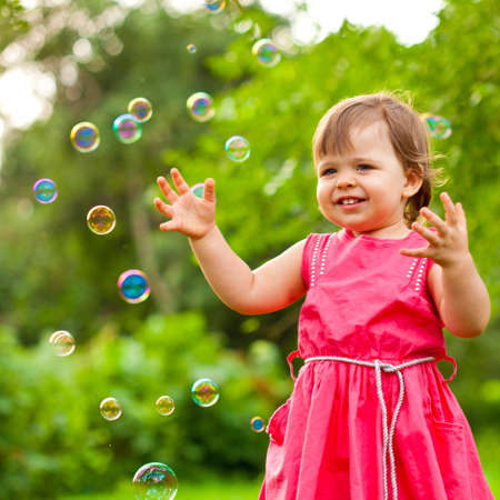 happy little girl in pink dress at park with flying soap bubblesの写真素材