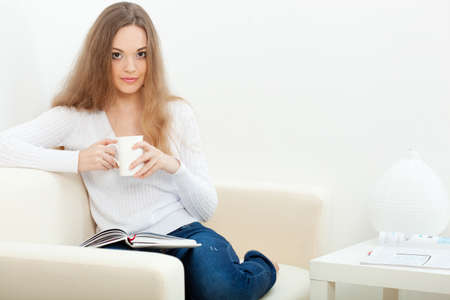 brunette  woman sitting on the sofa with white cup and reading the bookの写真素材