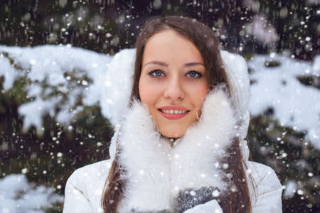 beautiful brunette woman standing under snowfall and holding her hands in mittens near faceの写真素材