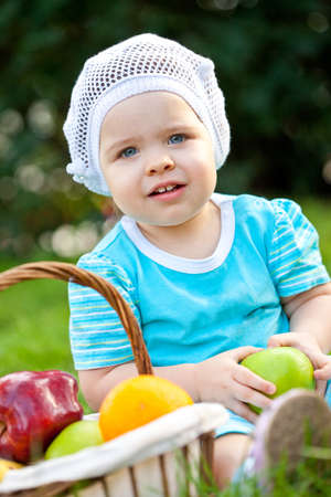 closeup portrait of baby girl sitting on the grass near basket with fruitsの写真素材