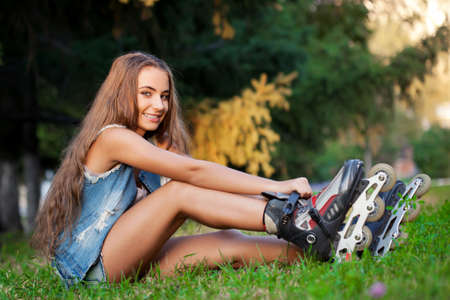 smiling girl sitting on the grass in park and puts on skatesの写真素材