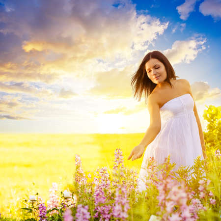 beautiful brunette woman walking in a field at sunset and touching flowersの写真素材