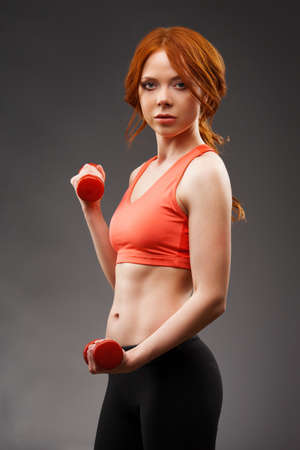 red -haired woman excersising with red dumbbells over gray and looking at cameraの写真素材