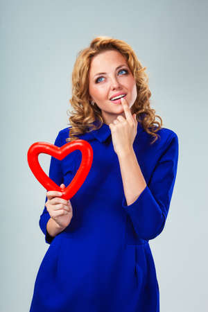 happy blonde woman wearing blue dress holding red heart symbol and thinkingの写真素材