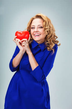 happy blonde woman wearing blue dress hugging red heart symbol , valentines day themeの写真素材