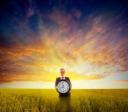businesswoman standing on field before the sunset and holding big clock