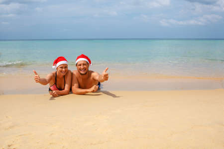 Happy romantic couple in red Santa hats at tropical beach relaxing on sun bedsの写真素材