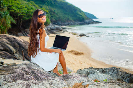 brunette woman sitting on the rock with laptop near  sea and looking to the cameraの写真素材