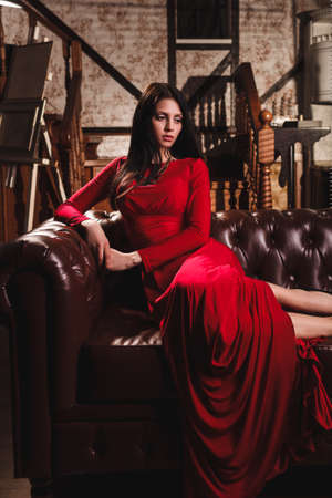 elegant sensual young brunette woman in red dress sitting on leather sofaの写真素材