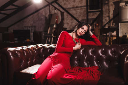elegant sensual young brunette woman in red dress sitting on leather sofaの写真素材