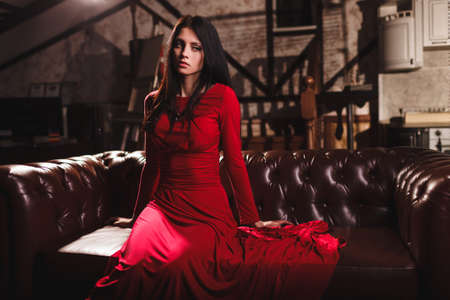 elegant sensual young brunette woman in red dress sitting on leather sofaの写真素材