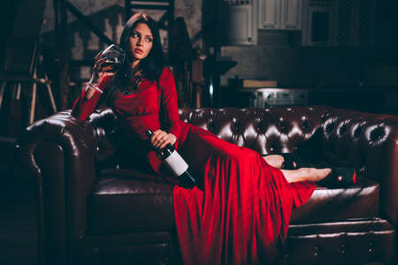 elegant sensual young brunette woman in red dress sitting on leather sofa, specially tonedの写真素材