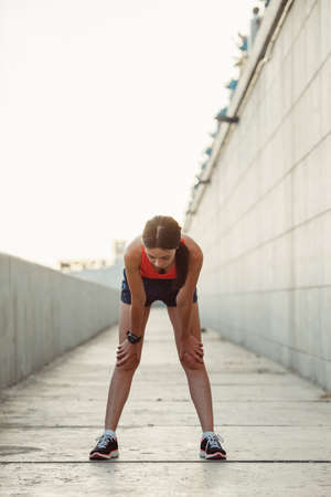 Young caucasian woman taking breath after jogging. Female athlete resting with hands on knees and looking down at sunsetの写真素材