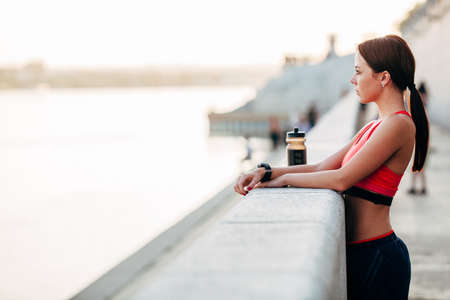 Female runner with bottled water tired from running standing near granite parapet, looking far awayの写真素材