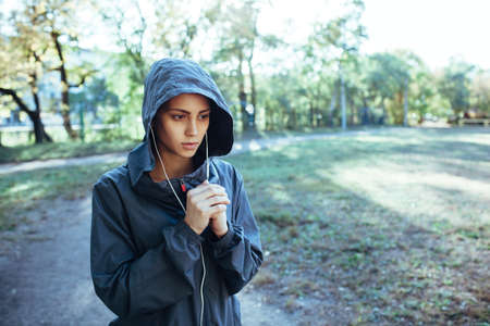 sporty woman wearing hood jacket and listening music outdoors in the park, she is warming her hands