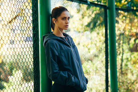 sporty woman wearing  jacket and listening music outdoors in the park, standing near mesh fence