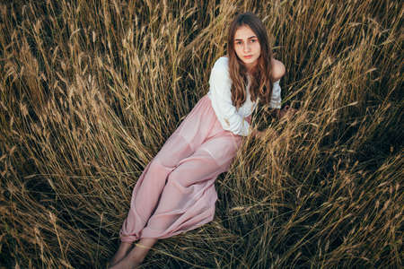 Young beautiful woman wearing white  and pink dress sitting in wheat fieldの写真素材