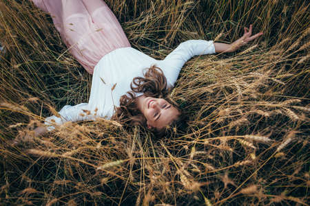 Young beautiful happy woman wearing white  and pink dress lying in wheat field, looking up