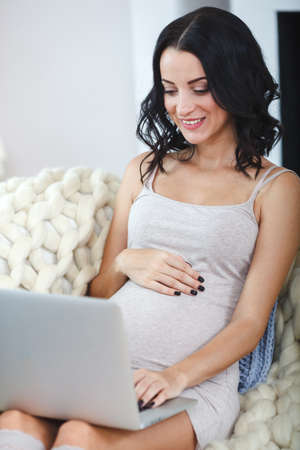 Beautiful young pregnant woman with laptop sitting on sofa at homeの写真素材