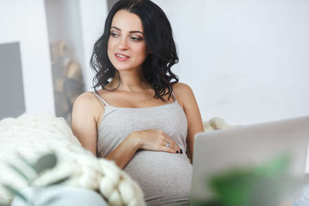Beautiful young pregnant woman with laptop sitting on sofa at homeの写真素材