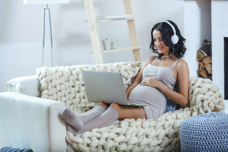 Beautiful young pregnant woman with laptop sitting on sofa at homeの写真素材
