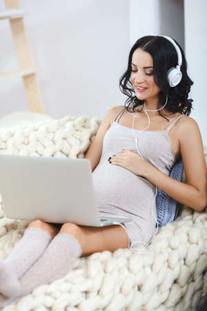 Beautiful young pregnant woman with laptop sitting on sofa at homeの写真素材