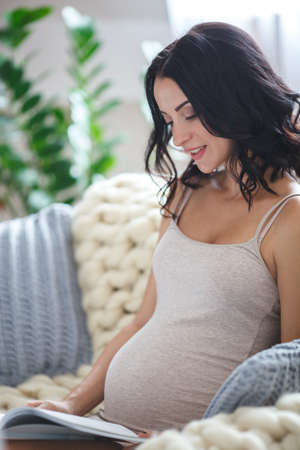smiling pregnant woman sitting on sofa and reading magazineの写真素材