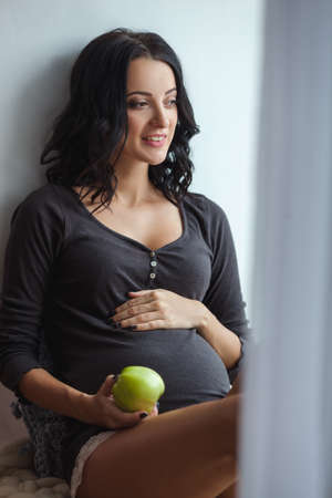 Cute happy pregnant woman on window sill with apples in the room, close upの写真素材
