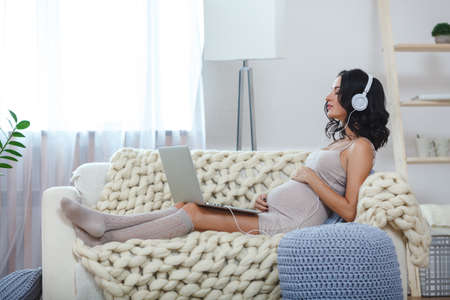Beautiful young pregnant woman with laptop sitting on sofa at homeの写真素材