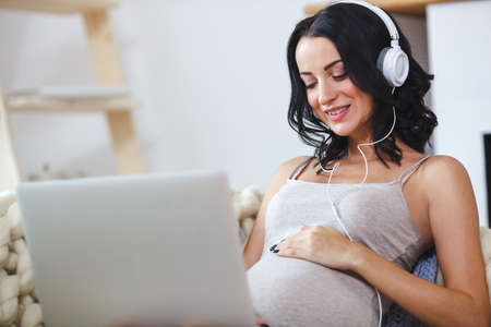 Beautiful young pregnant woman with laptop sitting on sofa at homeの写真素材