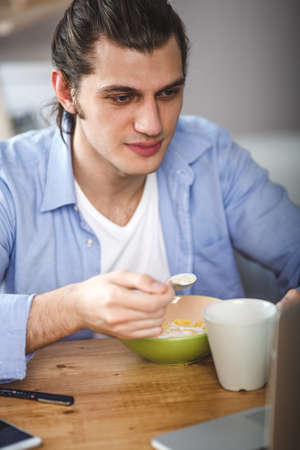 Young man eating corn flakes with milk and and looking to the laptop screenの写真素材