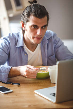 Young man eating corn flakes with milk and and looking to the laptop screen