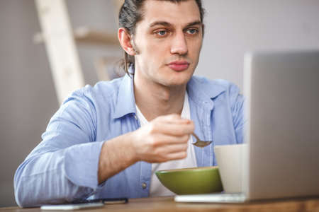 Young man eating corn flakes with milk and and looking to the laptop screenの写真素材
