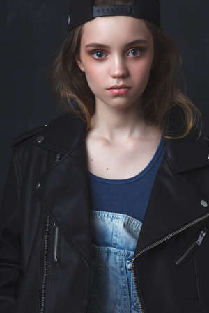 Young teenage girl wearing leather jacket jeans and cap, studio portrait over dark backgroundの写真素材