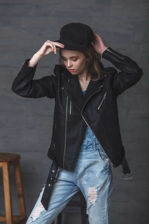 Young teenage blonde cute girl wearing leather jacket jeans and cap, studio portrait over dark backgroundの写真素材