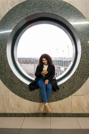 Young woman at the railway station sitting on round window and looking yo smartphoneの写真素材