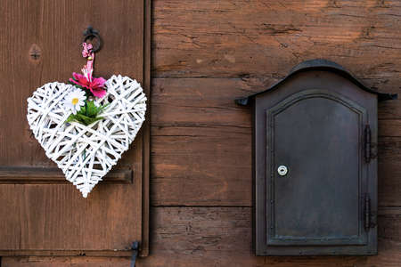 A wicker white heart with peonies and daisies hangs on a wooden shutter and a mailbox next to itの写真素材