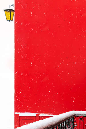View of a snowy balcony made of forged metal against the background of a red wall of a building during a snowfall and a street lamp. Concept architecture, winter, postcard, christmas, attention.の写真素材