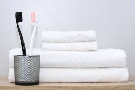 On a shelf in the bathroom are white towels of various sizes neatly folded and a glass cup with black and pink toothbrushes. The concept of morning procedures, order home, familyの写真素材