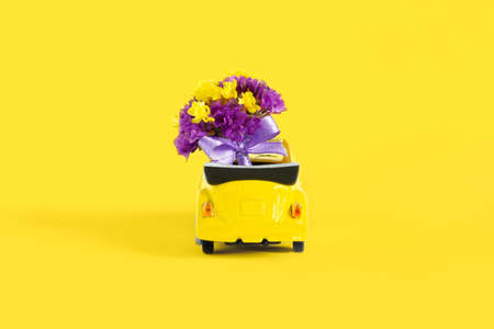 View of a colorful bouquet of purple flowers that is in a small yellow car on a yellow background. Selective focus. The concept of a holiday, wedding, flower delivery, giftの写真素材
