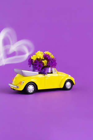 View of a yellow convertible car with a bouquet of flowers on a purple background behind which a train of a white heart develops. Concept holiday, delivery, art, background, transportの写真素材