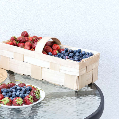 Fresh strawberries and blueberries lie in a plate and basket on a glass table against the background of a plastered white wall. Food concept, flat layout, gardening, summer, healthy eatingの写真素材