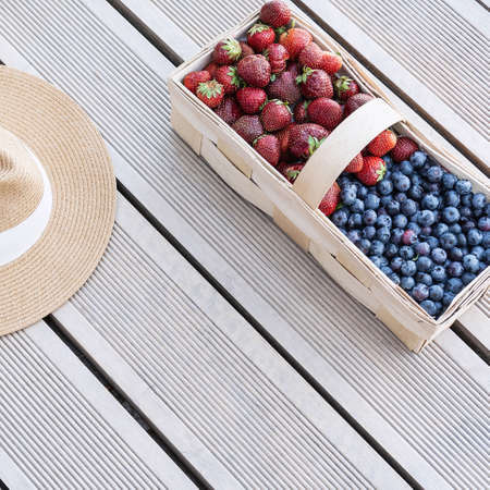 A hat and fresh berries of strawberries and blueberries lie in a basket on terrace boards on the veranda. Food concept, flat layout, gardening, summer, healthy eatingの写真素材