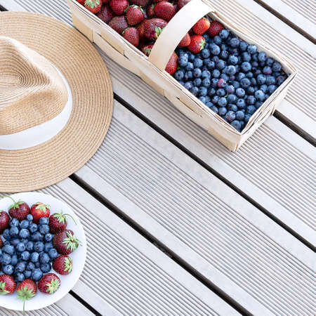 A hat and fresh strawberries and blueberries lie in a basket and on a plate on the terrace boards on the veranda. Food concept, flat layout, gardening, summer, healthy eatingの写真素材