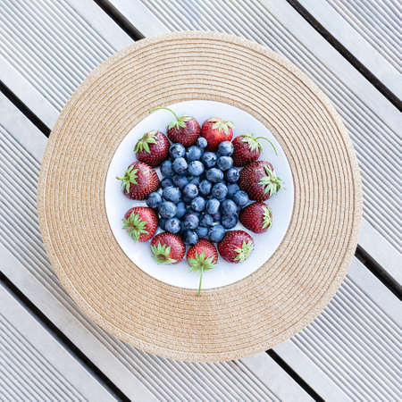Fresh strawberries and blueberries lie in a plate on a straw round substrate on terrace boards on the veranda. Food concept, flat layout, gardening, summer, healthy eatingの写真素材