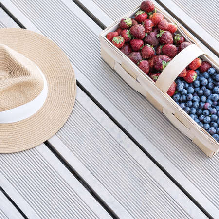 A hat and fresh berries of strawberries and blueberries lie in a basket on terrace boards on the veranda. Food concept, flat layout, gardening, summer, healthy eatingの写真素材