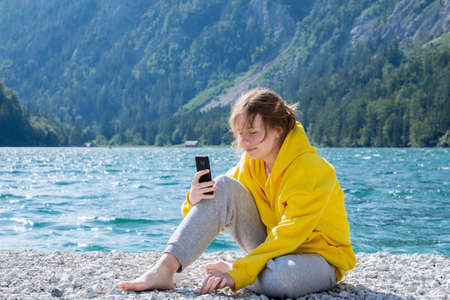 A girl in a bright yellow hoodie with a smile on her face sits on a lake in the mountains and looks at the smartphone. The concept of people traveling, communication, self-isolation.の写真素材