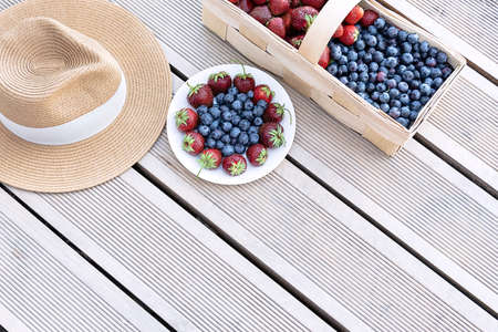 A hat and fresh strawberries and blueberries lie in a basket and on a plate on the terrace boards on the veranda. Food concept, flat layout, gardening, summer, healthy eating.の写真素材