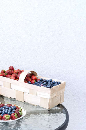 Fresh strawberries and blueberries lie in a plate and basket on a glass table against the background of a plastered white wall. Food concept, flat layout, gardening, summer, healthy eating.の写真素材