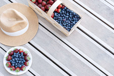 A hat and fresh strawberries and blueberries lie in a basket and on a plate on the terrace boards on the veranda. Food concept, flat layout, gardening, summer, healthy eatingの写真素材
